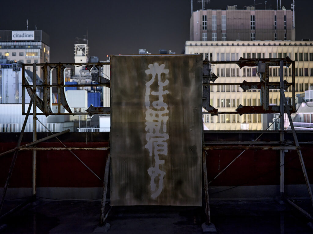 ＜5階以上＞ 「光は新宿より」2023, サイアノタイプ、看板,  夜の時間に視認できる、「奈落」の中から天空に伸びるサーチライトの光。向かって右手には、サイアノタイプ (青写真) 製の看板「光は新宿より」。終戦直後、焼け野原と化した新宿を地ならしし、電柱を建て、戦後初の闇市「新宿マーケット」を立ち上げた関東尾津組による、物資買取のための新聞広告と市場に掲出されたスローガンである。（撮影：森田兼次）