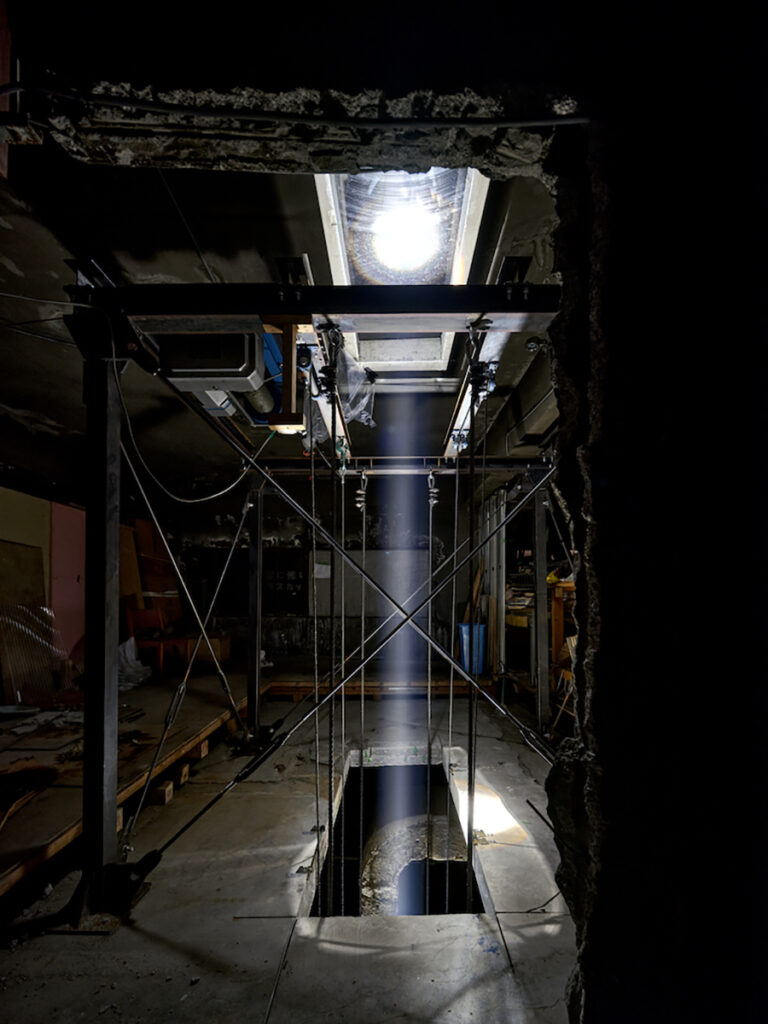  ＜2階＞「奈落」2023, サイトスペシフィック・インスタレーション, 歌舞伎町の名前が歌舞伎座誘致頓挫という戦後の都市計画に由来することに注目し、王城ビルで約30年間閉ざされてきた吹き抜け空間に「奈落」を設定。仏教用語で地獄を意味する「奈落」は、そこで働く人々がかつて「穴番」と呼ばれていたように、穴や沼のように仄暗く捉えられてきた。 本作では、空間に尾上右近の自主公演『研の會』による公演中の奈落で録音された歌舞伎公演の音を流し、カッティングした5階と屋上のスラブを「セリ」として上下に動かす。セリはサーチライトを搭載し、1時間に1回往復、夜になると上空に一筋の光を伸ばす。 さらに足元の2階の床（1階天井）も解体。閉鎖されていた「奈落」の概念を街と天へと上下左右に拡張する。（撮影：森田兼次）