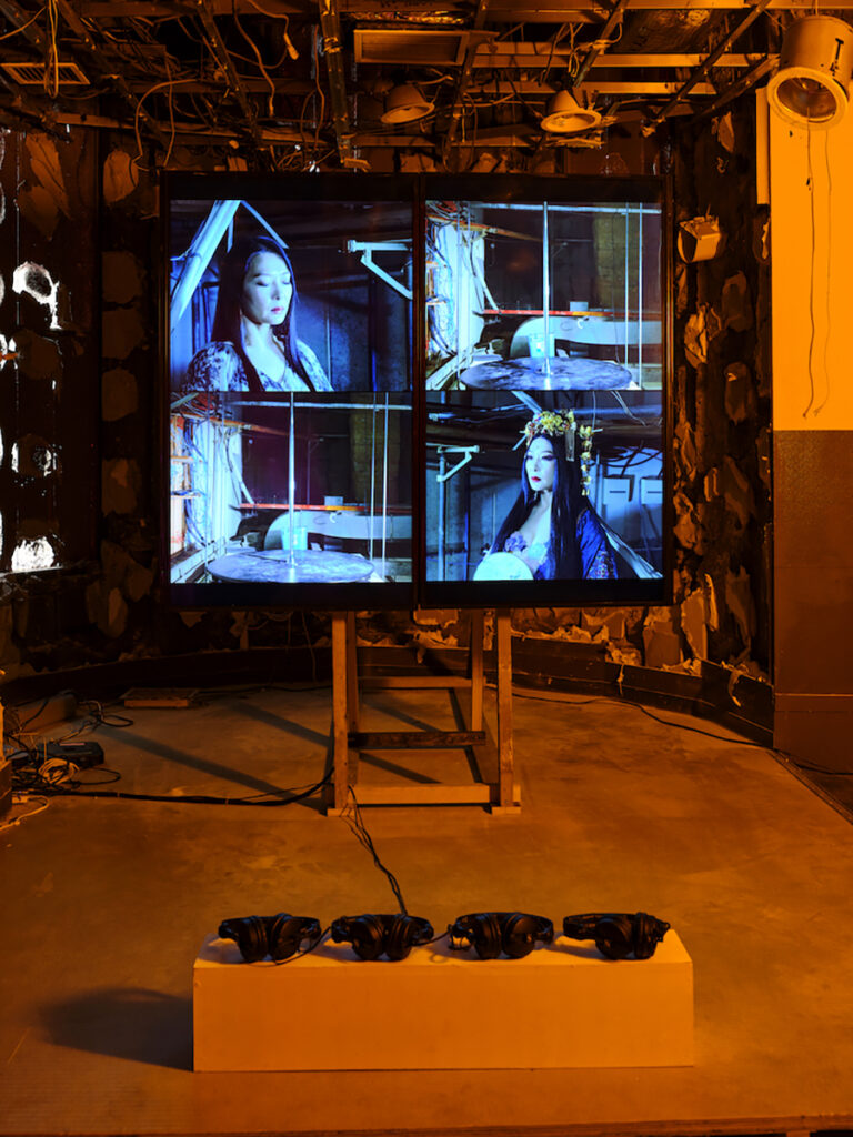 ＜2階＞「I（アイ）」2023, Chim↑Pom from Smappa!Group＋KUMI＋コンスタン・ヴォアザン, ビデオインスタレーション,  本展で弁財天を演じるのは、日本屈指のポールダンサーKUMIである。弁財天として、高村月とともに吉原、歌舞伎町、日本橋でパフォーマンスをしてきたが、その土地土地にも弁天が祀られていたことに縁を感じてきたという。「私」を超越した神を演じるKUMIだが、その根底には在日朝鮮人3世としての「私」への葛藤があった。東京オリンピック2020の前夜祭に出演した際にも、国家事業と自身の立場の関係を問い続けたという。その難しさと最も向き合ったのは、東京ゲゲゲイがDANCE@HEROで優勝した際の作品「世界の果て」のサポートとして、劇中の「君が代」で踊った時だった。主宰のMIKEYとの信頼関係に基づくその行為は後のKUMIに多大な影響を与えたが、今回、再び「君が代」で踊るアイデアが持ち上がったことで葛藤が再来。Chim↑Pomと日本在住のフランス人映像作家コンスタン・ヴォアザンと会議を重ねながら、撮影日前日まで相談を続ける。当日、「奈落」で「君が代」が流れるなか、無人のポールを見つめるKUMIと弁財天KUMI両者の表情が撮影された。（撮影：森田兼次）