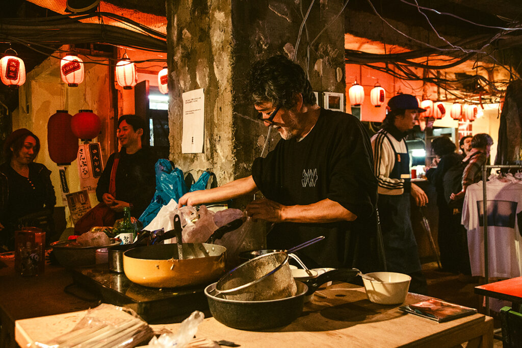 プログラム：王城ビル(A)-⑤アー横｜再開発ブームで増え続ける東京の横丁に対するオルタナティブとして、アーティストやアートコミュニティが集まり、22:00から朝まで王城ビルで飲食やパフォーマンス、物販を展開する横丁イベントが開催される。ゆるく楽しめる場を目指し、「出来る範囲」が理想とされているため、売り切れや閉店が早い可能性はあるが、逆にダラダラと朝まで過ごせる見込みもある。基本的には美味しいものや楽しいことにこだわる作家たちが出店を予定しており、独自の魅力を持つイベントになるだろう。 深夜枠「アー横」では、会田誠が重慶の友人からもらった火鍋を日本人向けにマイルドにアレンジした特製火鍋を提供するほか、途中でやめる(山下陽光)が服を通じた金儲けをお題に客のマッチングを図る「即実験ランド」を開店する。また、Chim↑Pom from Smappa!Groupの「金三昧」が、歌舞伎町最奥地のセレクトショップ「THE FOUR-EYED」とコラボしFAKE商品をリリース。金三昧ではカルト的人気のクリエイター「ぼく脳」の特製ドリンクも登場する。 大久保に存在したアートスペースUGOからは、UGO実行委員会が「UGOの給湯室」として白湯（概念）を提供。「とりま、白湯飲んで一息つこー」を合言葉に、歌舞伎町の喧騒にオアシスを出現させる予定だ。松戸のオフラインサロン・ノーウェアでは「ノイズカレー」と「コンドームおにぎり」が提供されつつ、Day2には意味不明なパフォーマーたちがブッキングされ、特撮ショーからパンク、ノイズ、ヒップホップまで、幅広いジャンルのライブが行われることになる。 さらに、墨田区の社会実装型アートスペース「京島駅」も神輿的な展示とタンドール窯で焼く「宇宙のパワーナン」を提供し、札幌からも、札幌国際芸術祭のカウンター展「すすきの夜のトリエンナーレ」が、「北海道・すすきの物産展」として参加。日本各地の多様なカルチャーとコミュニティが交錯する、アーティスト横丁が2夜に渡って立ち現れる。（撮影：上原俊）