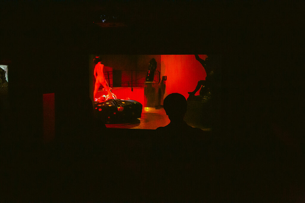 プログラム：王城ビル(A)-⑥遠藤麻衣『オメガとアルファのリチュアル』｜本作は、2024年に国立西洋美術館で開催された展覧会「ここは未来のアーティストたちが眠る部屋となりえてきたか？」のために、エドヴァルド・ムンクの《アルファとオメガ》に触発されて制作された映像作品である。ムンクの異種交配の世界観を背景に、国立西洋美術館でのヌードパフォーマンスと日本のストリップ劇場の歴史を重ね合わせた内容で、現役ストリッパー宇佐美なつと遠藤自身が共演する。 本作は、西洋美術史と日本の戦後ストリップ文化をフェミニズムの視点から批評し、女性の身体性に焦点を当てている。歓楽街において身体の問題は今も根深いものとして存在するが、一方で、歌舞伎座招致の計画が頓挫し、その名前だけが残った歌舞伎町での展示はまた特有の文脈も強調する。新宿の帝都座の額縁ショウを起源とする戦後のストリップ文化、遊郭から広まった歌舞伎など歴史的文脈を踏まえ、芸術と芸能の境界を再考させる。（撮影：上原俊）