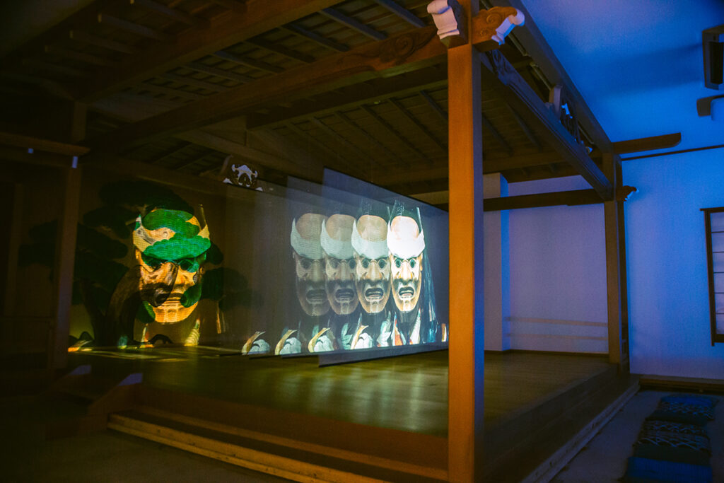 プログラム：新宿歌舞伎町能舞台(C)-①渡辺志桜里｜渡辺志桜里、安田登、加藤眞悟、ドミニク・チェンによる新作能「射留魔川」が、安原杏子 a.k.a 青椒肉絲の映像インスタレーションとして登場。「射留魔川」は「人間宣言」をテーマに、天皇制神話や単一民族の幻想からの解放、多様性と自然との共存を問い直す作品です。近代以降、生態系の象徴から人間中心主義の象徴とされた天皇が、敗戦後ヒトとしての姿に戻るも、依然として一般的な人間像とは異なる公的存在であることを描写しています。 また、インスタレーションには歴代天皇の肖像がガラスの面として制作される渡辺の新シリーズも展示される予定です。（撮影：上原俊）